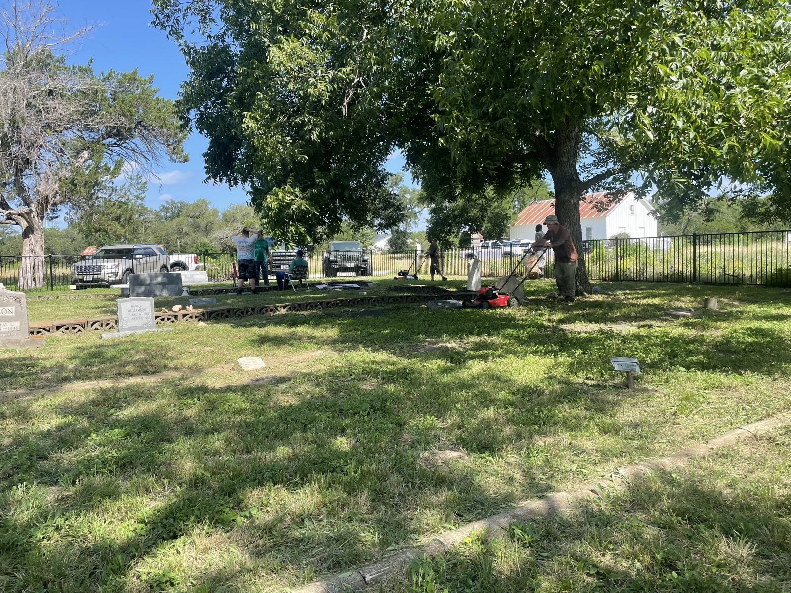 Several volunteers with push mowers work among the headstones under large pecan trees, the white Nameless Schoolhouse visible in the distance.