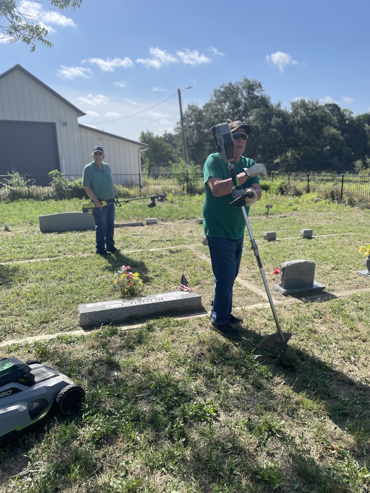 A volunteer in a face shield uses a string trimmer near headstones while another works in the background.