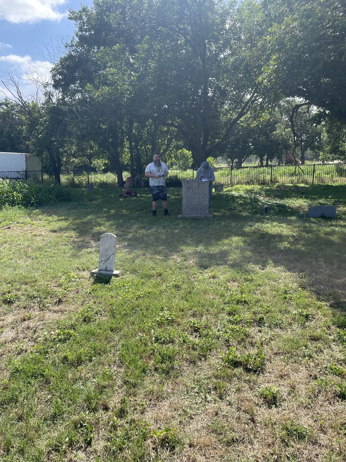Two volunteers stand near a tall stone monument with a mower parked at the fence line behind them.